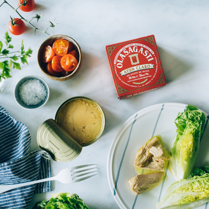 Packaging of Yellowfin Tuna - Steak in Brine, from Olasagasti. A package is open, and you can see the inside. Dish with lettuce and Yellowfin Tuna - Steak in Brine. Bowl with chopped tomato and bowl with salt.