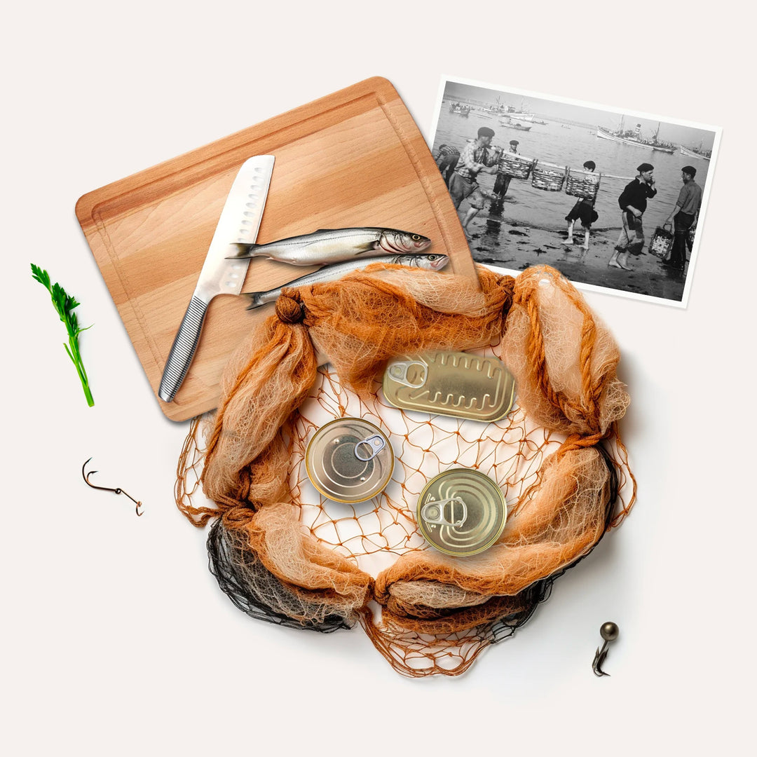 Illustrative image of our history: fishing net, cans of canned goods, hook, old black and white photograph of fishermen unloading their fish onto the beach in baskets, wooden board, two sardines and knife.