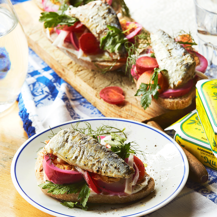 Sardines in Olive Oil served on a plate on a slice of bread, with tomato and fresh vegetables. Wooden board with two more slices of bread with preserved sardines and fresh vegetables.