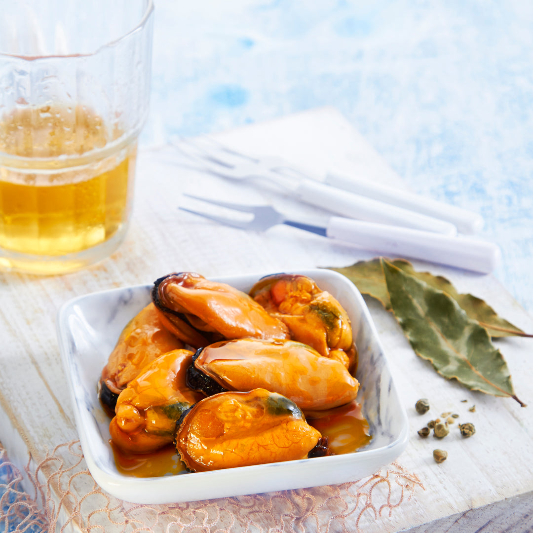 Plated serving of Los Peperetes Giant Mussels in Escabeche. Large, plump, and vibrant orange Galician mussels (PDO) served in a white dish. The background features a cold beer and laurel leaves, showcasing the premium texture and artisanal quality of this traditional Spanish seafood appetizer.